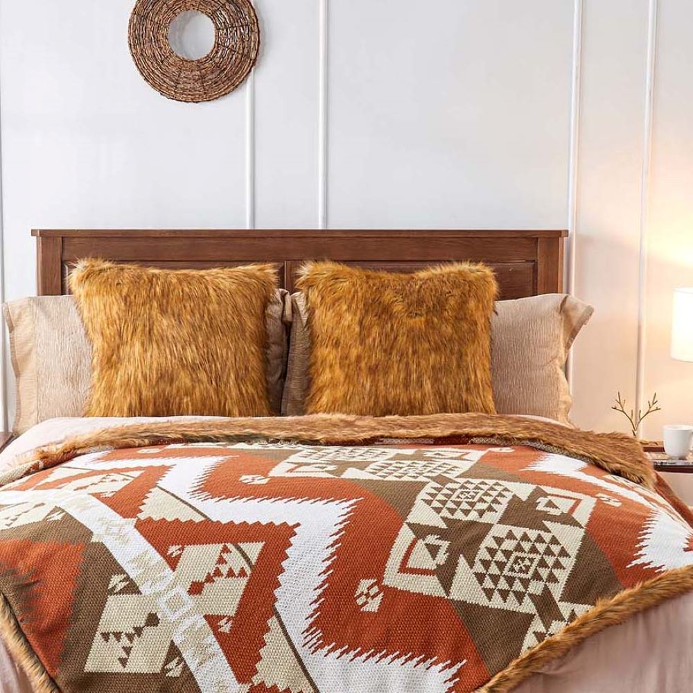 A cozy bedroom setup featuring a wooden bed with two fluffy orange pillows and a colorful blanket with geometric patterns in shades of orange, brown, and white.