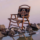 Wooden chair with Aztec-patterned fabric seat and backrest on rocky lakeshore at sunset.