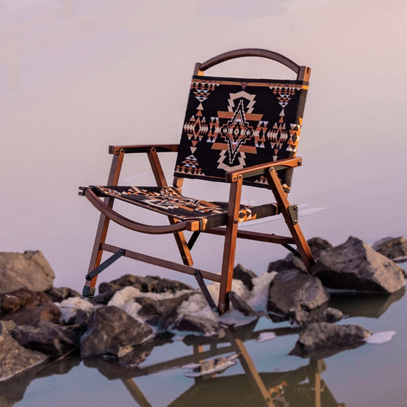 Wooden chair with Aztec-patterned fabric seat and backrest on rocky lakeshore at sunset.