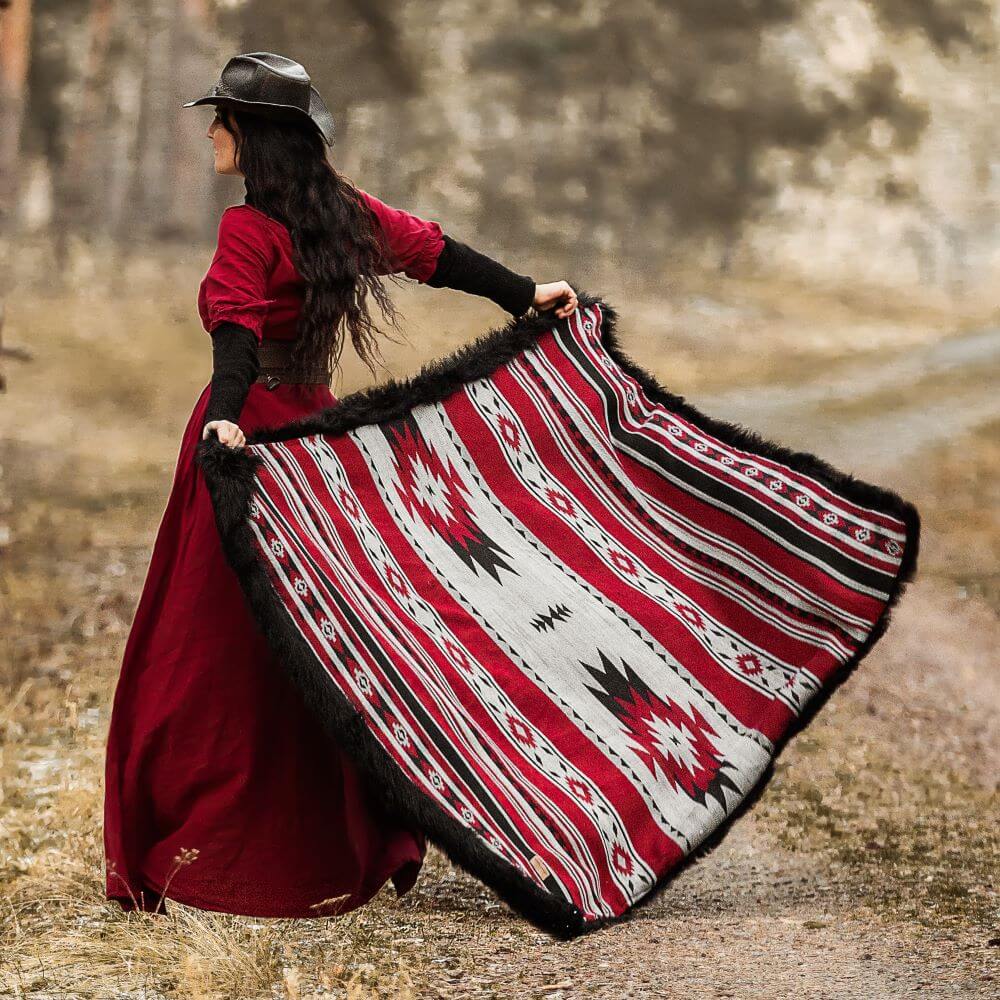 A woman in a red dress and black hat holding a large, colorful patterned blanket with red, white, and black geometric designs in an outdoor setting.