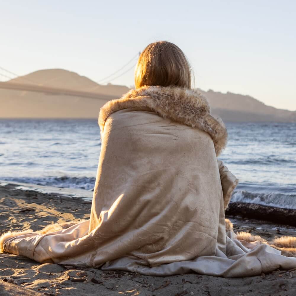A blonde woman sits on the beach by the Golden Gate Bridge, wrapped in a champagne-colored leopard faux fur blanket looking out at the sea.