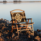 Wooden chair with Aztec-patterned fabric seat and backrest on rocky lakeshore at sunset.