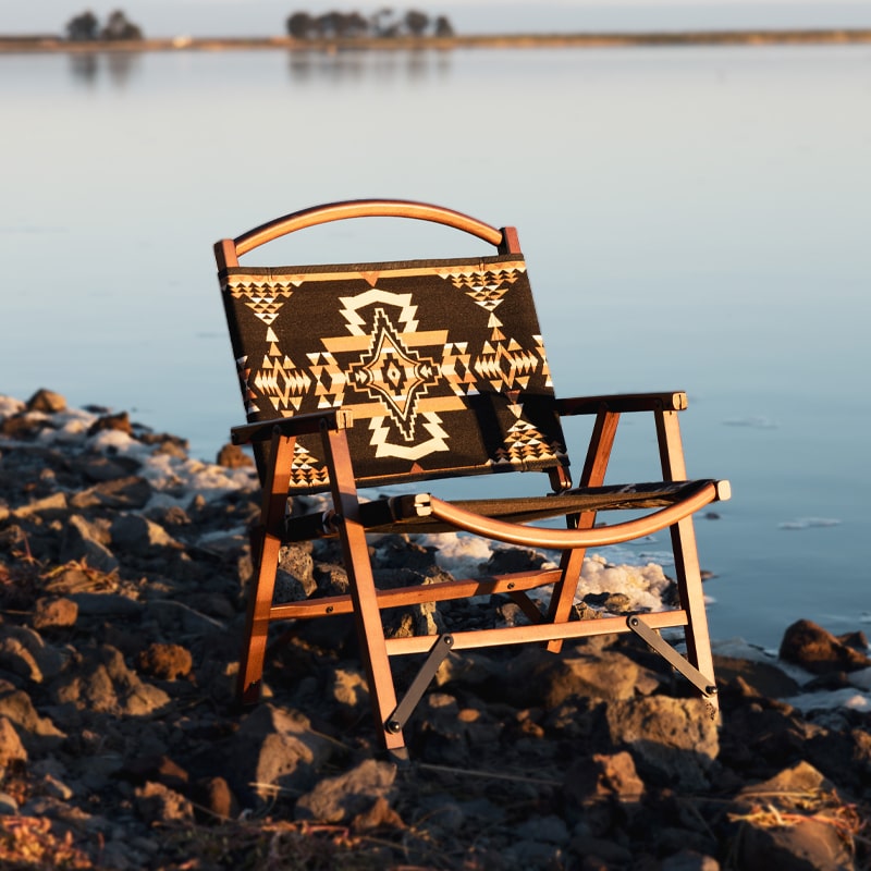 Wooden chair with Aztec-patterned fabric seat and backrest on rocky lakeshore at sunset.