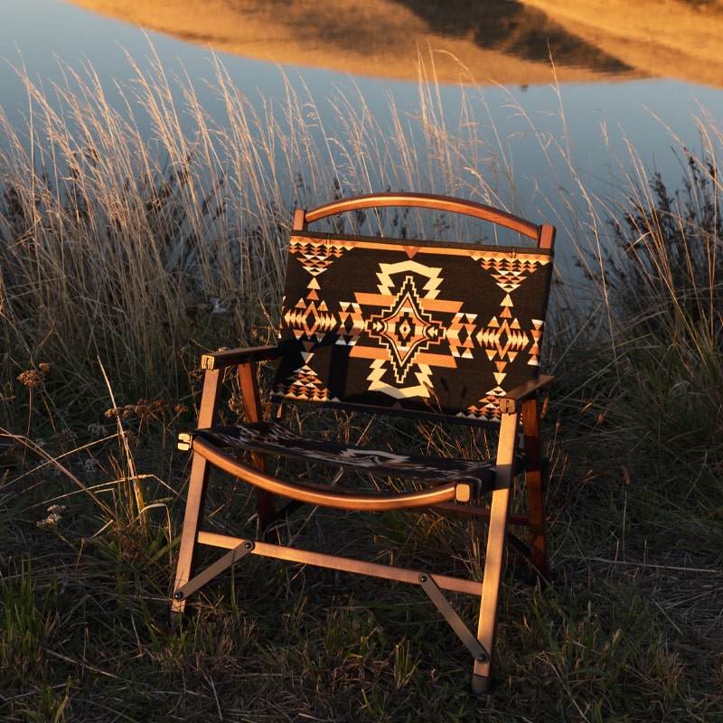 Wooden camping chair with Native American-inspired geometric pattern set against a scenic backdrop of rolling hills and a lake.