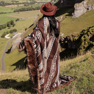 A woman in a brown hat wrapped in a large, brown and beige patterned blanket with geometric designs, standing on a grassy hill overlooking a scenic valley.