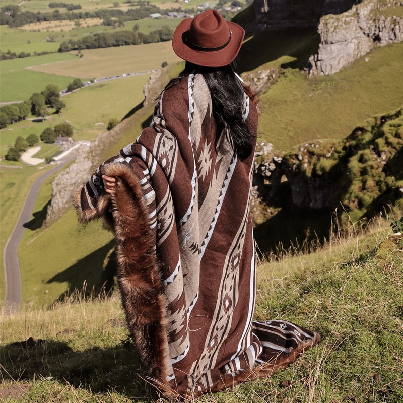 A woman in a brown hat wrapped in a large, brown and beige patterned blanket with geometric designs, standing on a grassy hill overlooking a scenic valley.