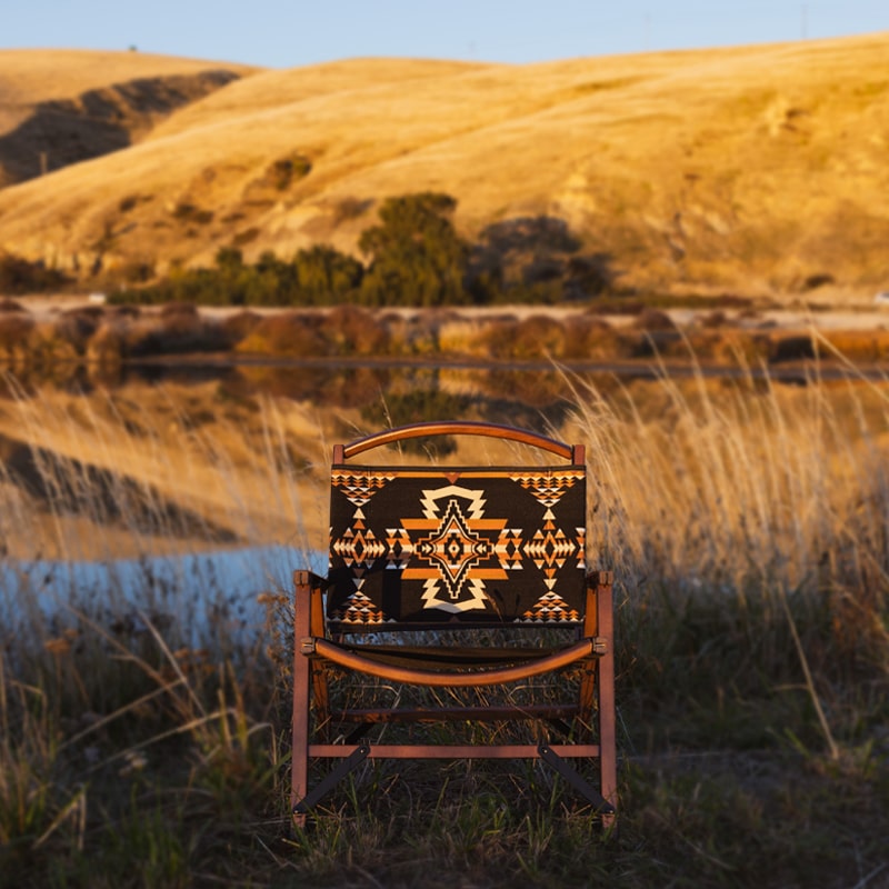 Wooden camping chair with Native American-inspired geometric pattern set against a scenic backdrop of rolling hills and a lake.