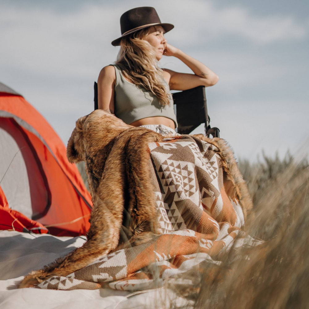 A person wearing a hat and sitting on a camping chair next to a red tent, wrapped in a patterned blanket and looking into the distance.