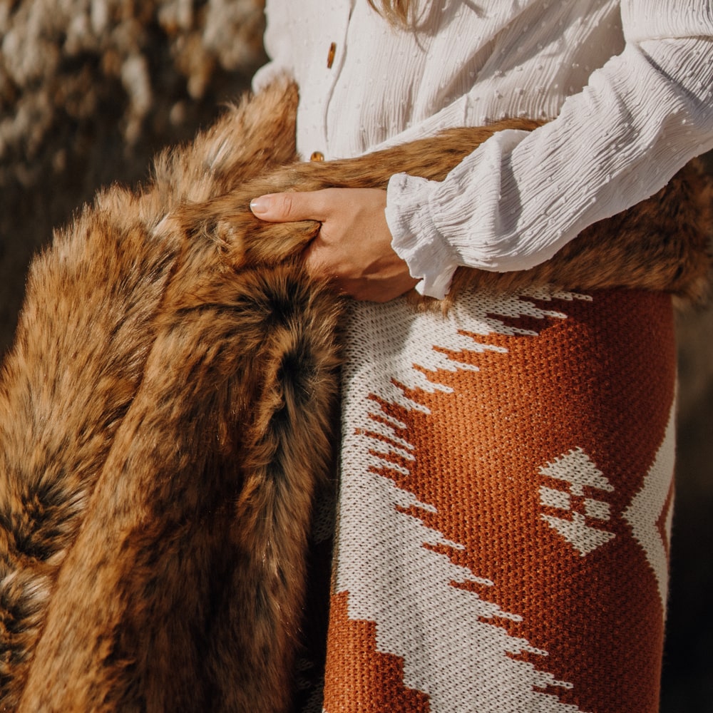 A woman wearing a white shirt holding a fur-lined, patterned blanket wrapped around their waist.