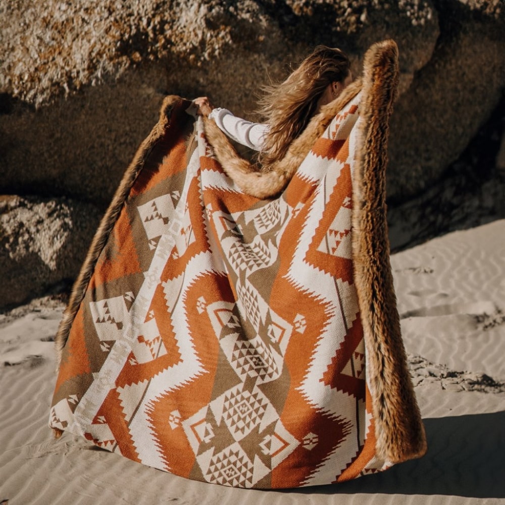 Patterned alpaca wool blanket with geometric design in shades of orange, brown, and white, draped over a rock on sandy ground, reflecting cultural textile art.