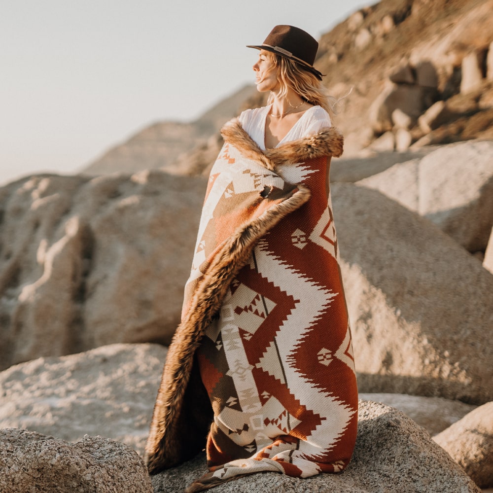 Woman wear a har and is wrapped in a patterned blanket with warm tones, standing on rocky coast.