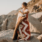 Woman wear a har and is wrapped in a patterned blanket with warm tones, standing on rocky coast.