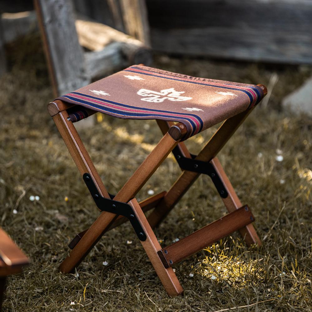 Anifurry wooden outdoor stool with a beech wood frame and a seat with a Thunderbird-inspired woven pattern, folded and placed on grass.