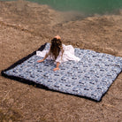 Woman lying on a blue and white patterned faux fur blanket beside a lake, wearing a white dress.