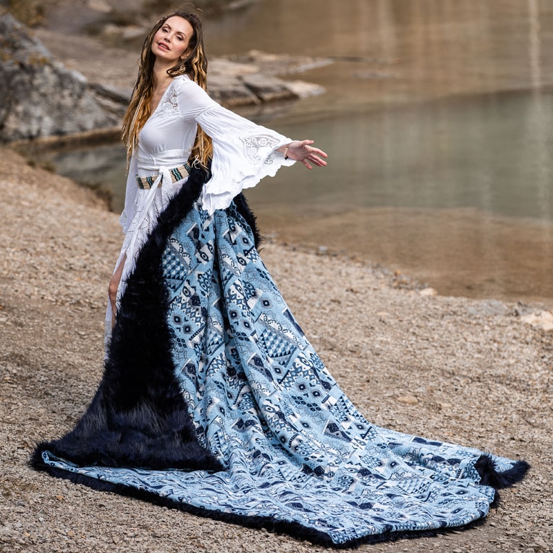 Woman standing outdoors in a long, flowing dress with a white upper part and blue patterned design towards the bottom, ending in black fur-like material at the hem, against a natural backdrop with rocks and water.