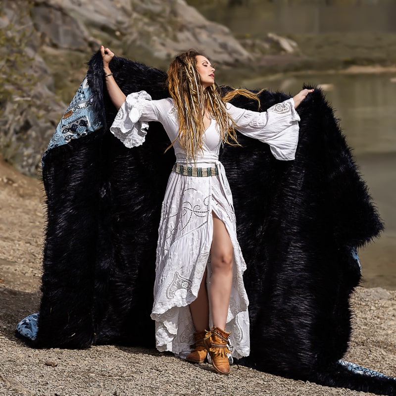 Woman standing outdoors holding up a large black cloak with blue lining, wearing a white patterned dress, brown boots, and standing against a backdrop of rocky terrain and water body.