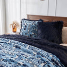 Neatly made bed with patterned blue and white comforter, various textured pillows including a black furry one, and a fluffy tan decorative item on the bedside table.