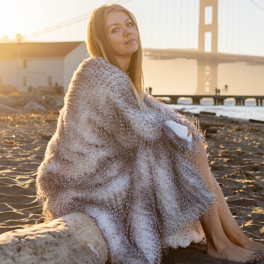 A beautiful woman sits with a gray porcupine faux fur blanket covered on the beach of the Golden Gate Bridge when the sun sets.