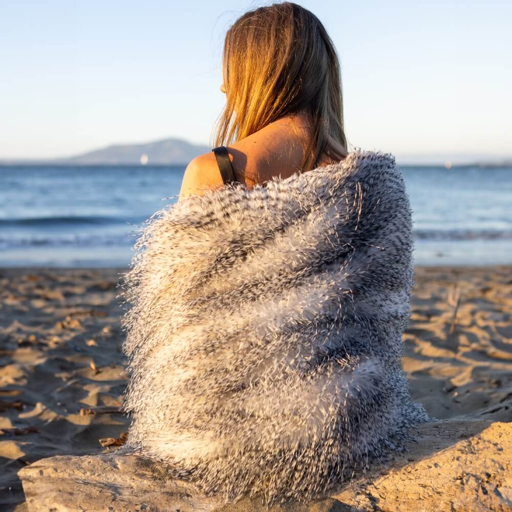 A lady cover with a porcupine faux fur blanket was sitting on the beach by the river.