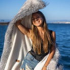 A beautiful woman is holding a a reversible blanket with one side porcupine faux fur and the other side in plush velvet with Anifurry trademark label on and smiling at the beach.