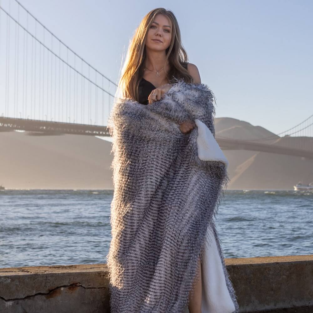 A blonde woman wrapped in a reversible grey porcupine blanket stood by the river at the Golden Gate Bridge.