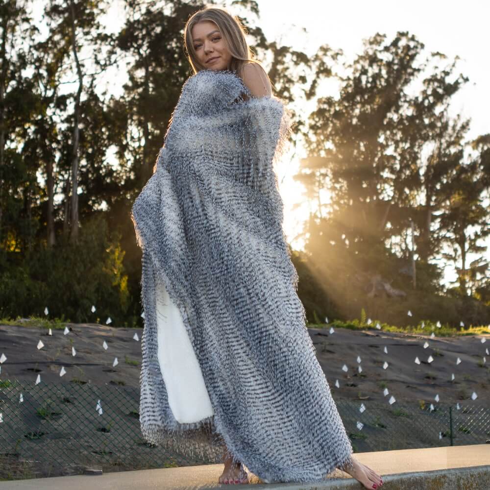 A beautiful woman covered with a gray porcupine faux fur blanket on the ground when the sun sets.