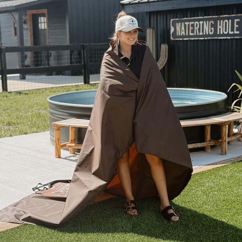 A woman standing on the grass, wrapped in an anifurry outdoor brown picnic blanket, accompanied by a water tank