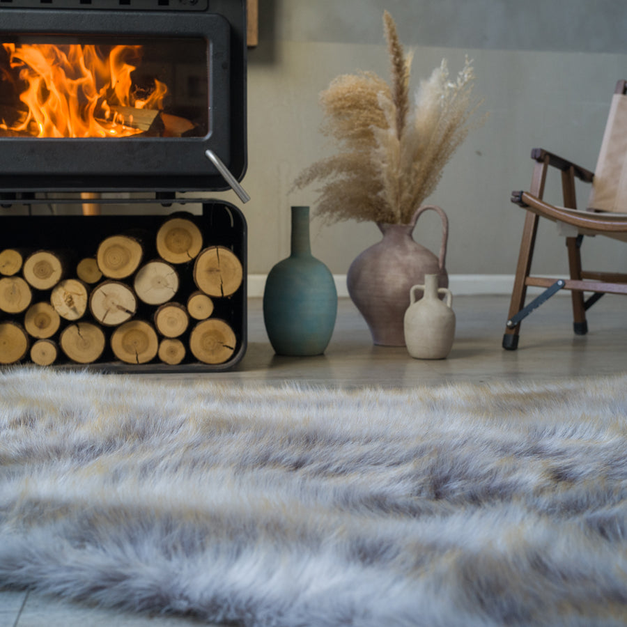 The grey fake fur rug is beside a fireplace. The rich-textured rug looks extremely soft and cozy. It's very warm and cozy.