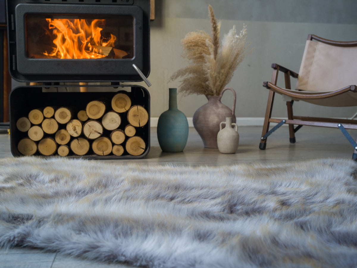 The grey fake fur rug is beside a fireplace. The rich-textured rug looks extremely soft and cozy. It's very warm and cozy.